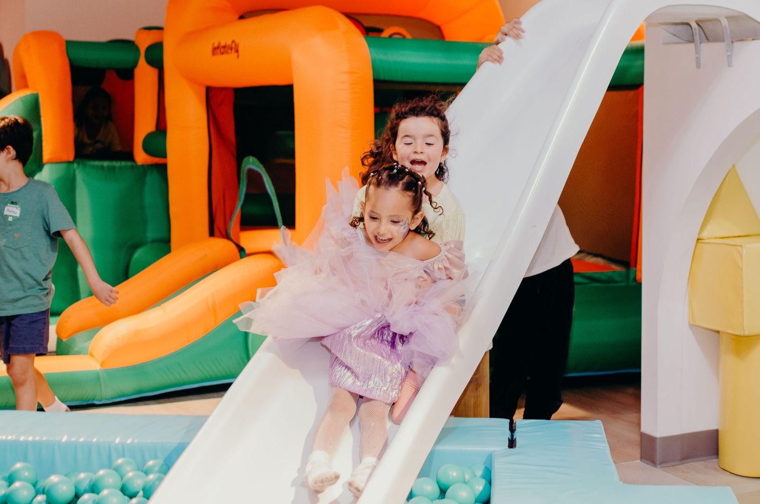 Two children having fun on inflatable slides and bounce houses at an indoor play area with colorful playground equipment.