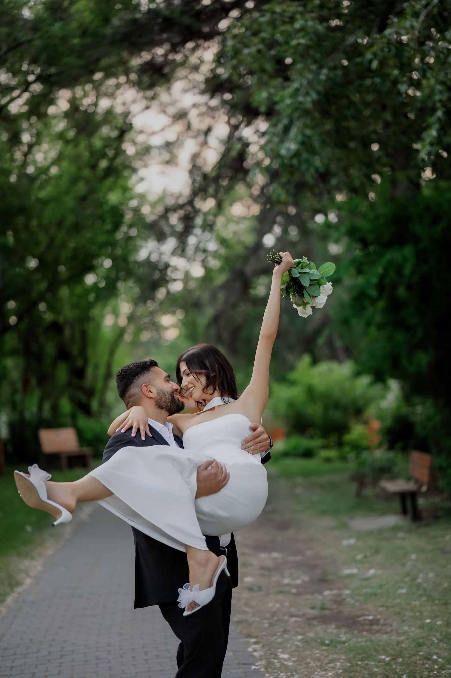 A romantic wedding couple embracing in a park setting with lush greenery and a tree-lined path in the background.