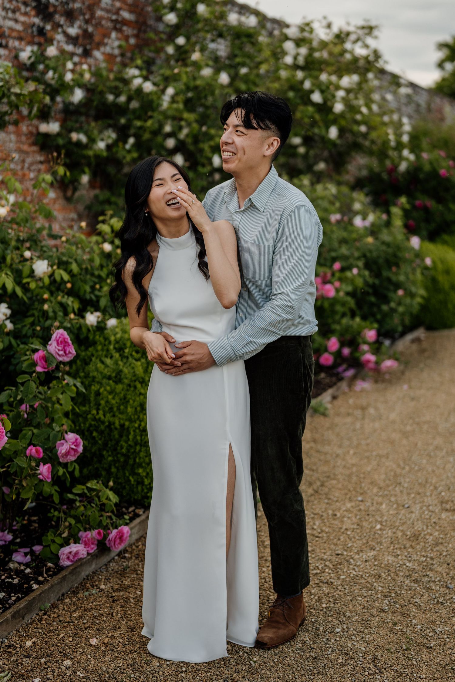 A couple embraces in a romantic garden filled with pink roses, the woman in a white gown and man in a blue shirt.