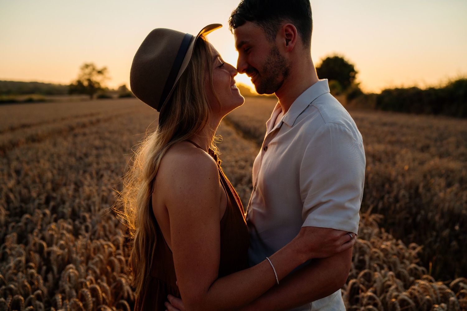 Couple sharing a romantic moment at golden hour in a wheat field, woman in hat and man embracing tenderly.