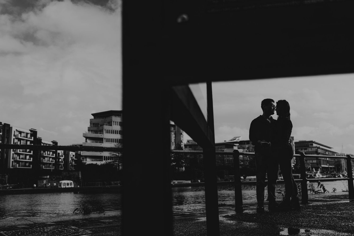 Romantic silhouette of a couple kissing by a waterfront with city buildings in a black and white photo.