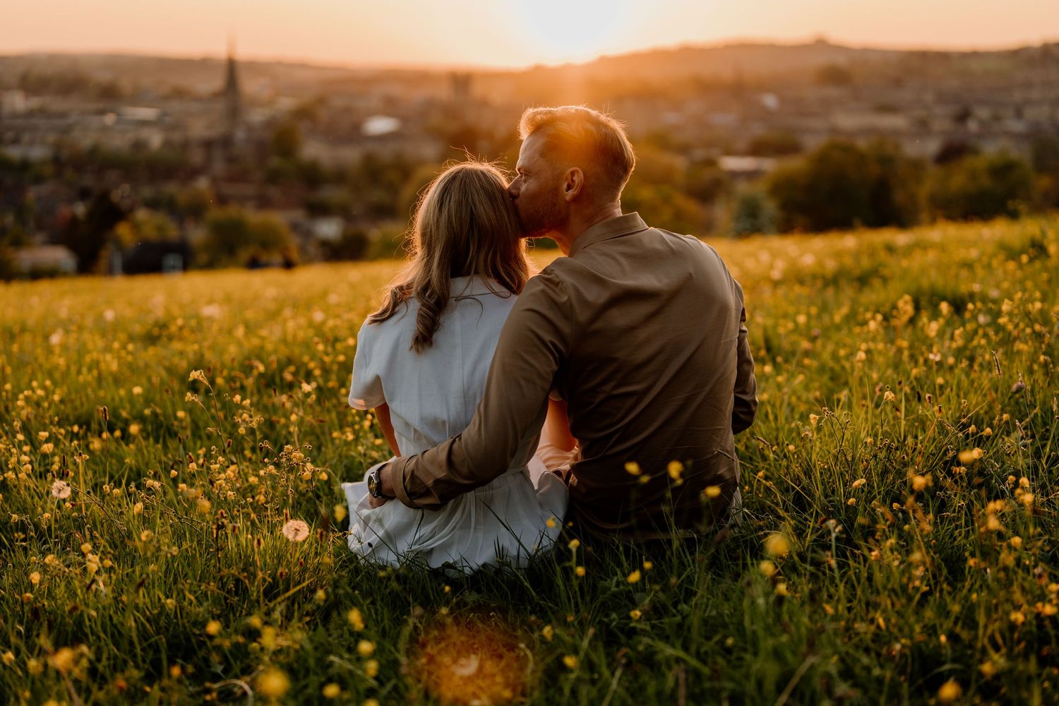 Couple sitting together in a sunny meadow of wildflowers watching a golden sunset over a city skyline.