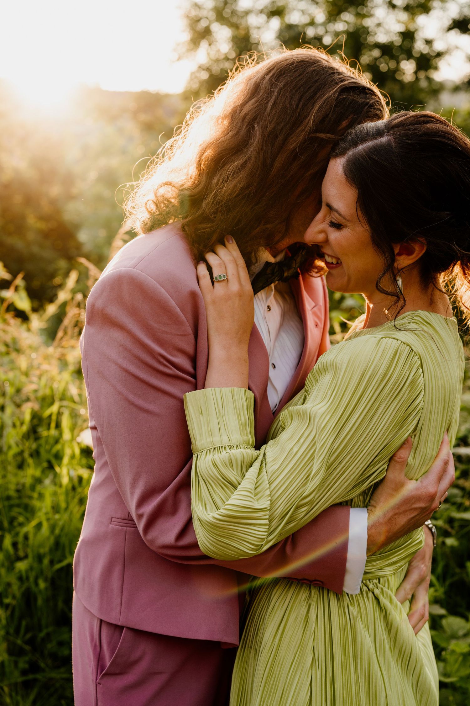 Couple embracing at golden hour, man in pink suit and woman in green dress laughing together outdoors.