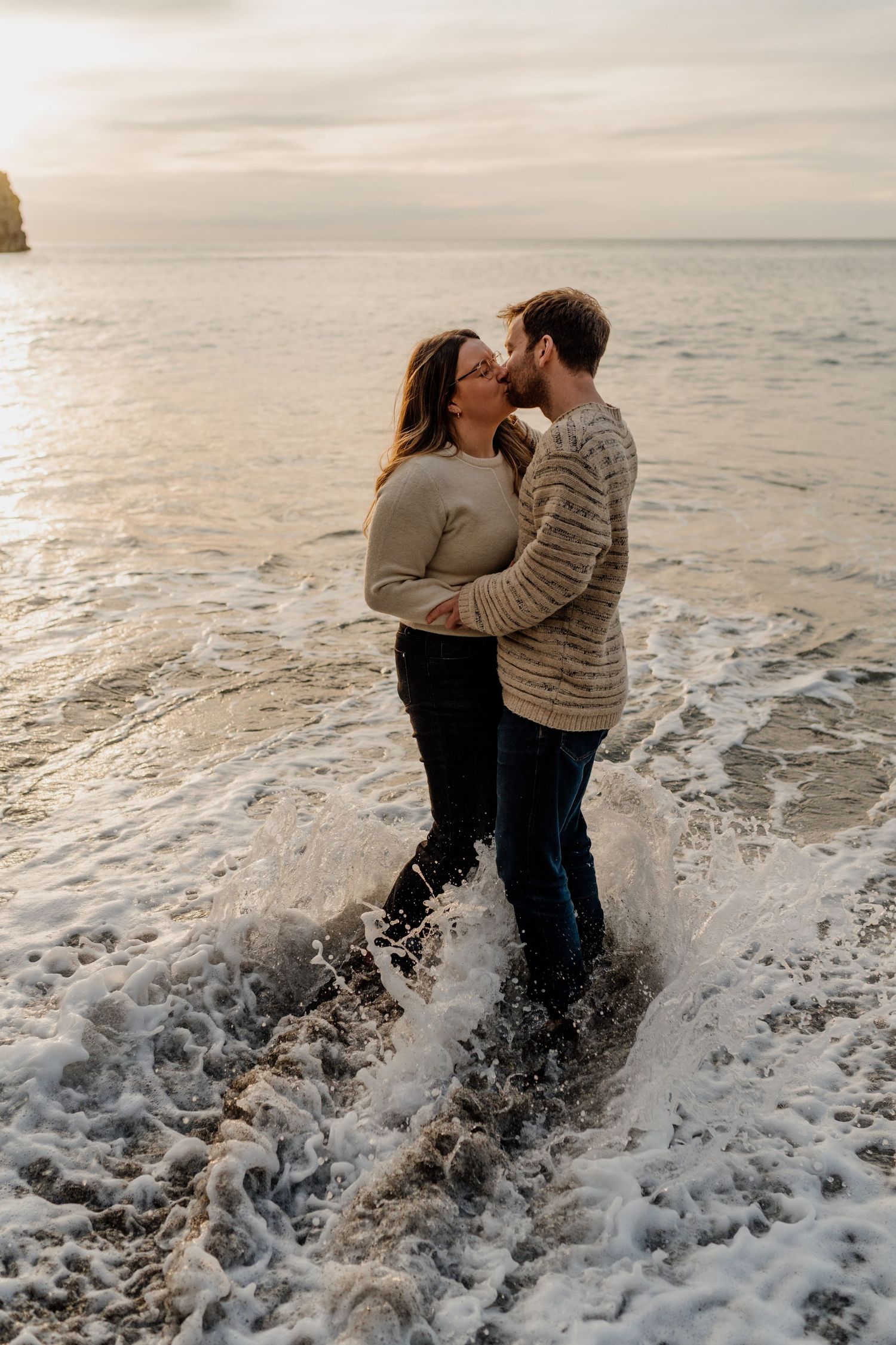 Couple sharing a romantic kiss while standing in ocean waves at sunset on a rocky beach.
