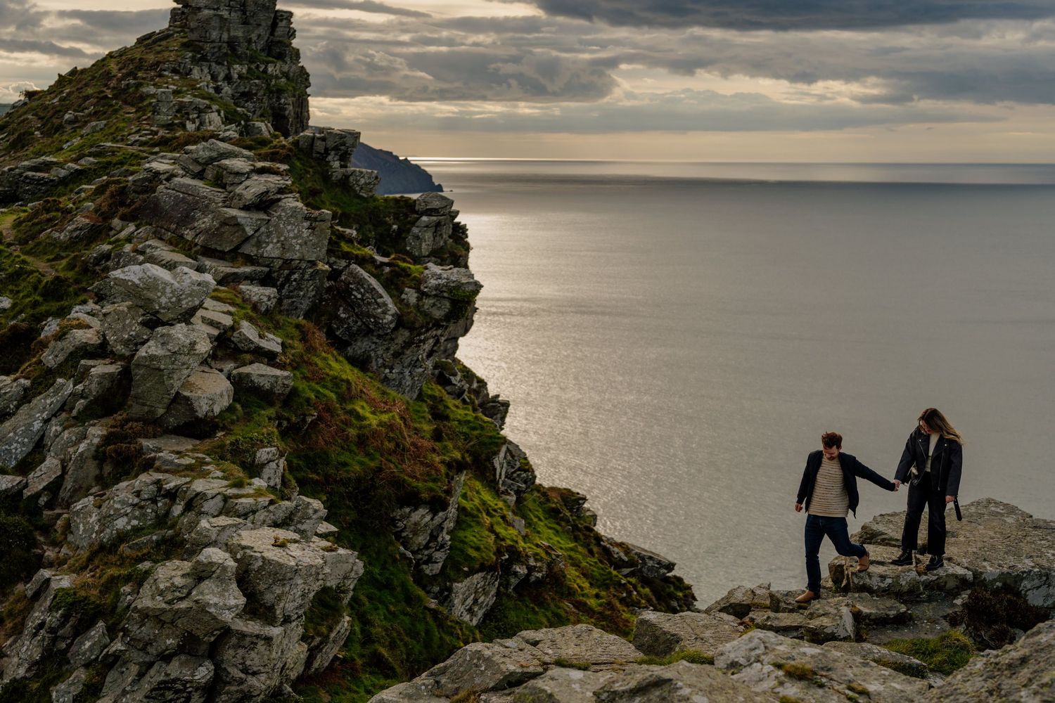 Couple holding hands while hiking on rocky coastal cliffs with dramatic ocean views and stormy skies.