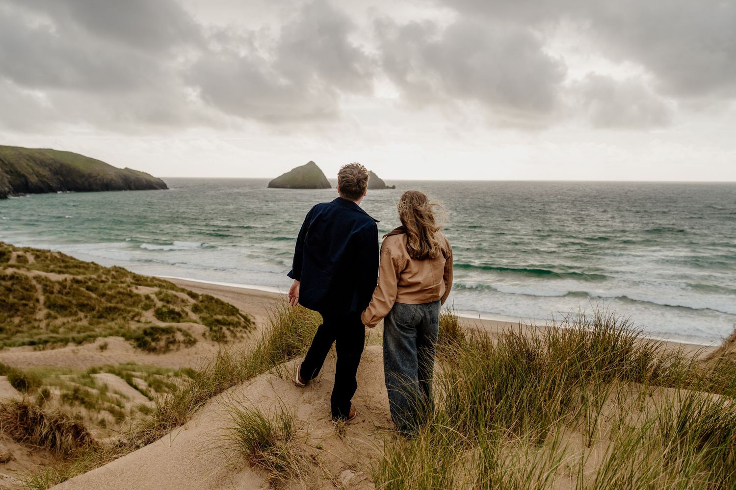 Couple holding hands walking on sandy beach dunes overlooking dramatic ocean coastline on overcast day.