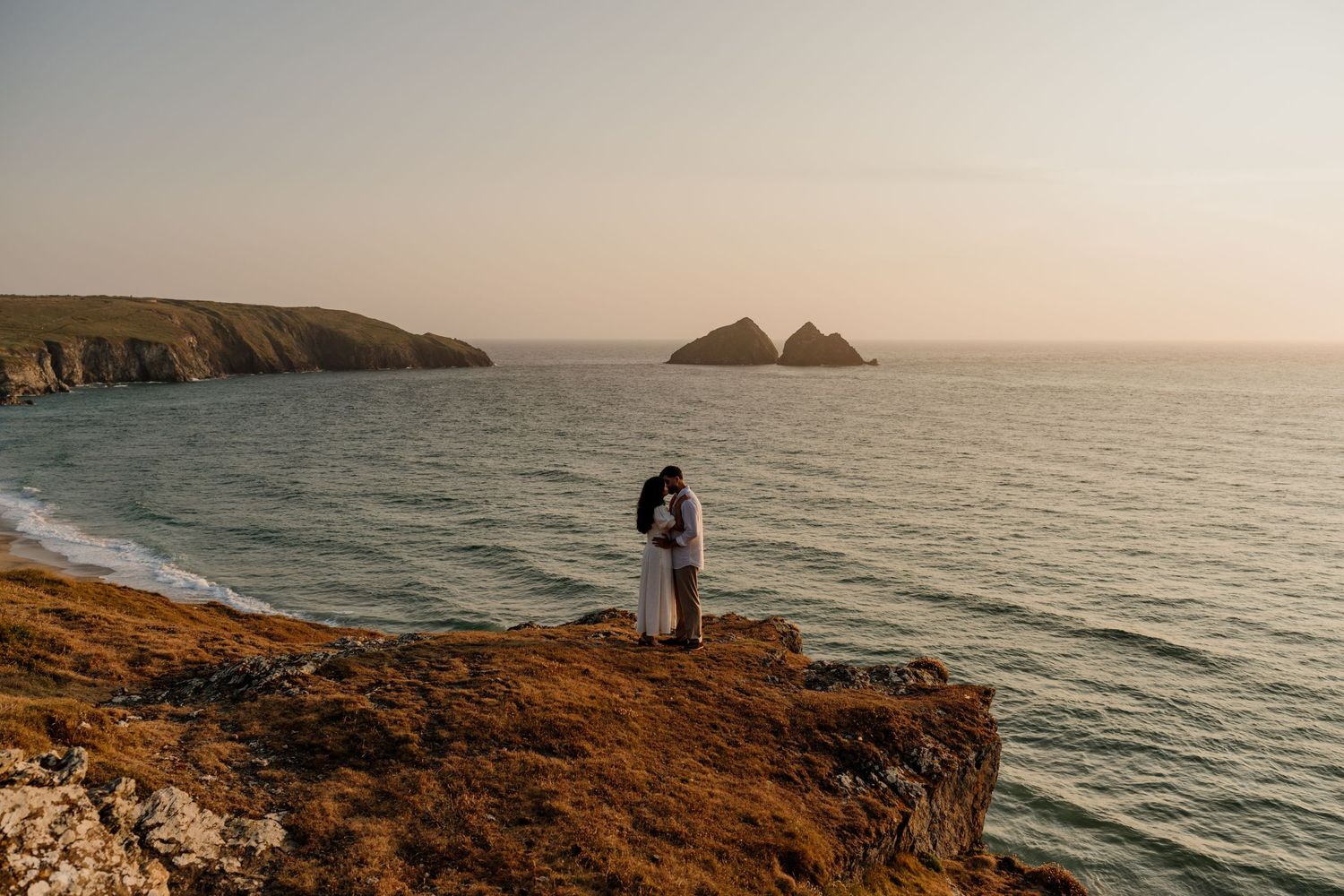 Couple embracing on a coastal cliff at sunset with rocky sea stacks and calm ocean in the background.