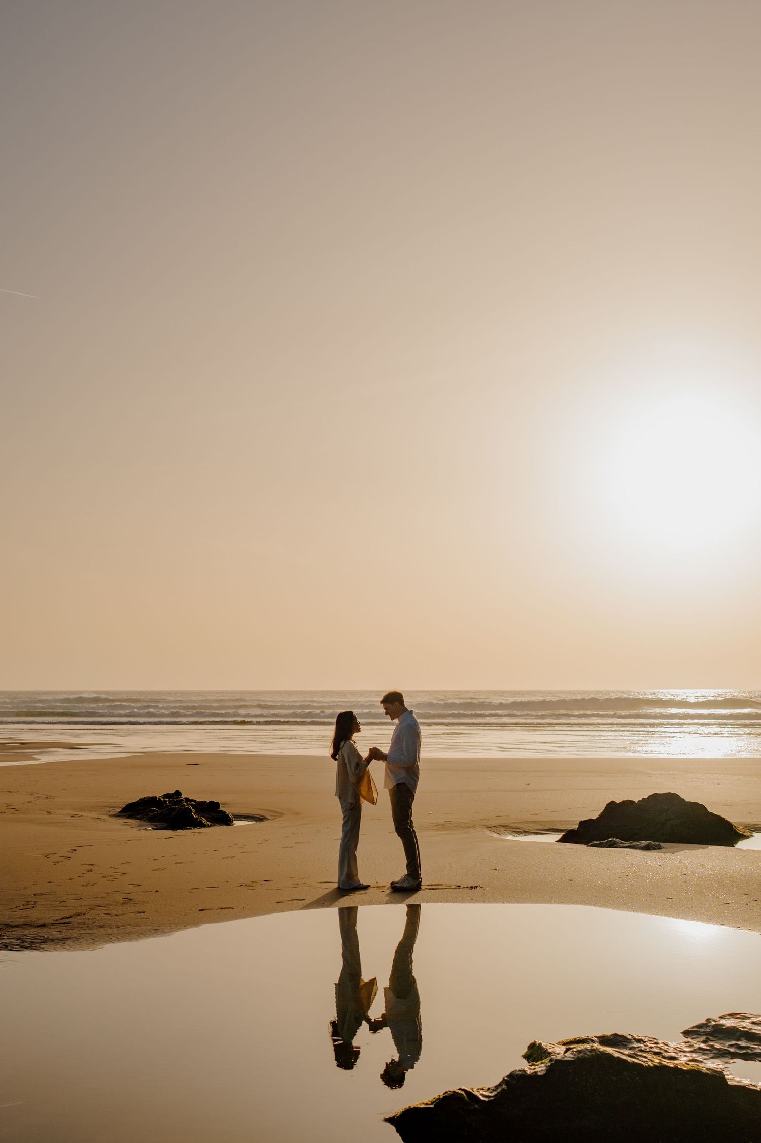 Couple holding hands on a sandy beach at golden sunset, with their reflection visible in the wet sand near the ocean.