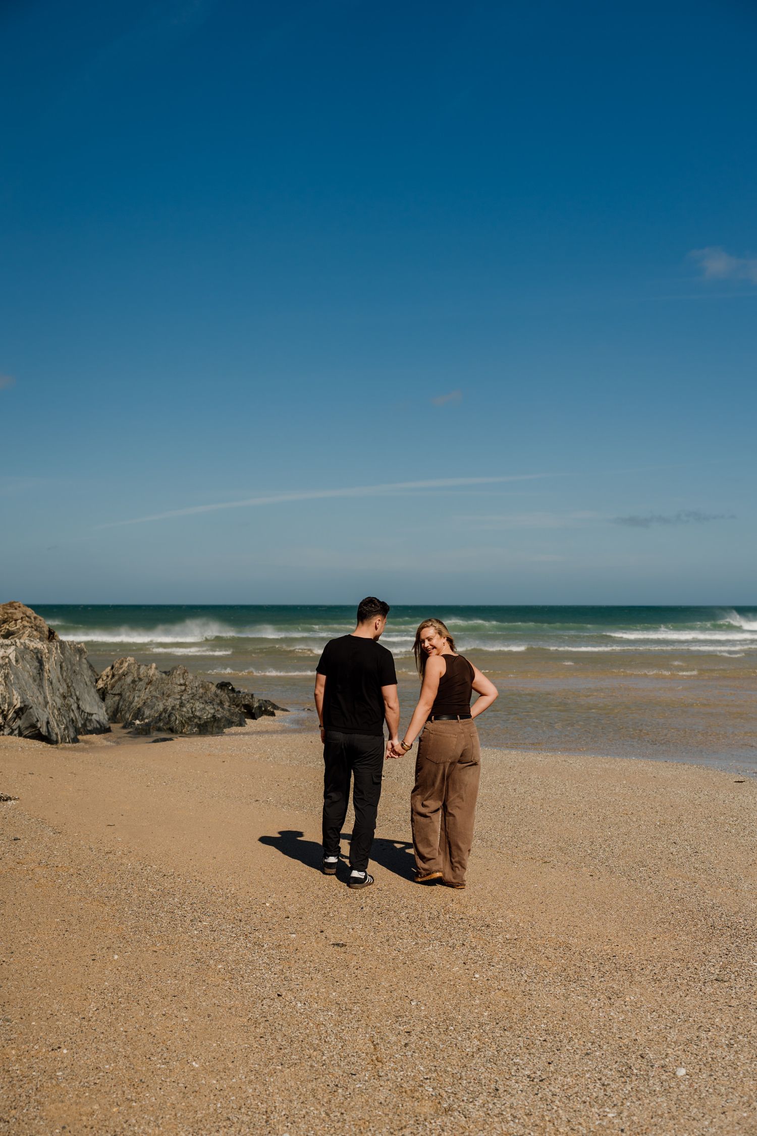 Couple holding hands on a sandy beach with ocean waves and blue sky backdrop.