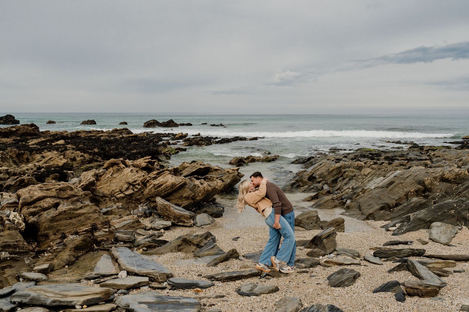 Couple embracing romantically on a rocky beach with ocean waves and cloudy sky in the background.