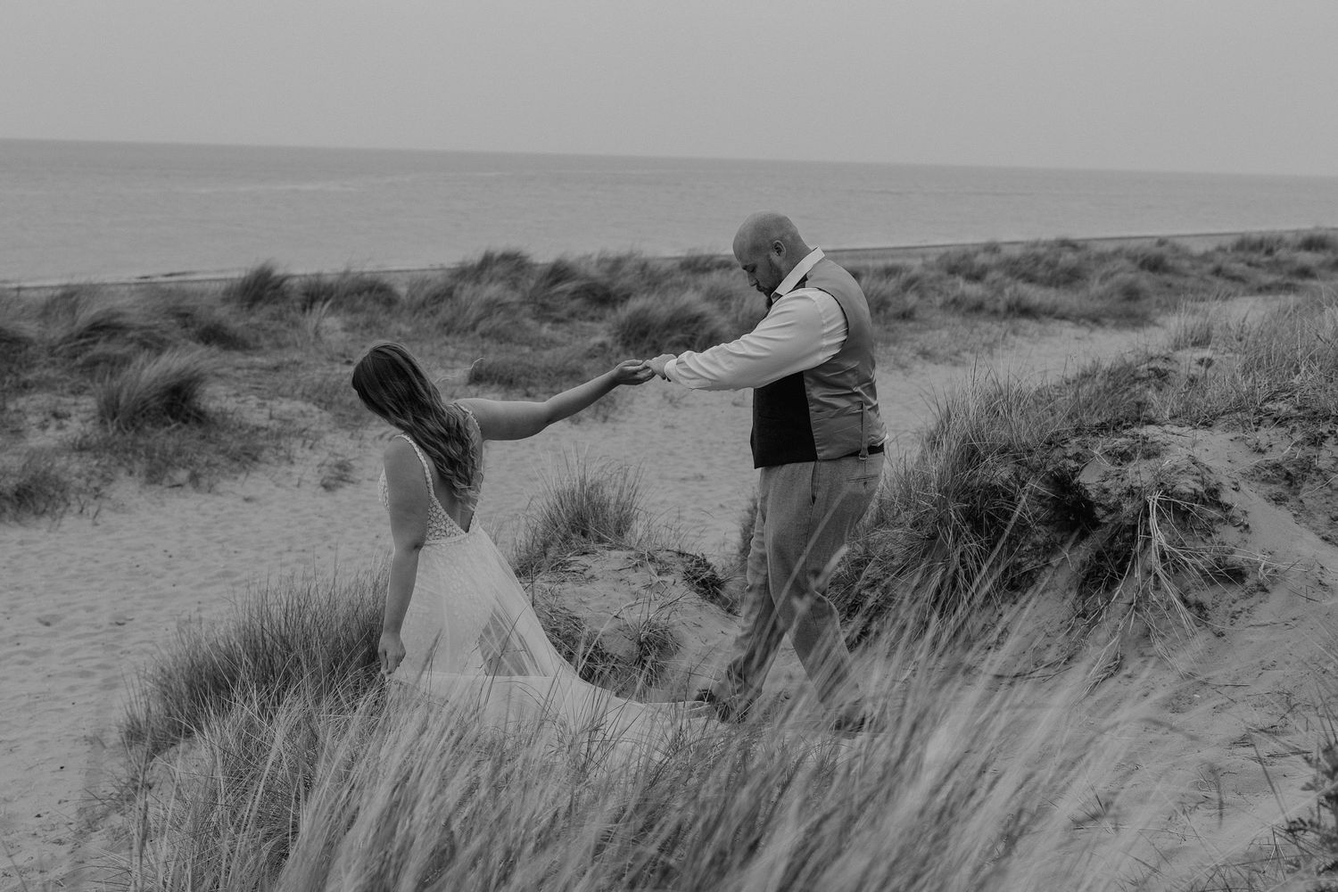 a bride is leading her husband but the hand down the sand dunes at the beach.