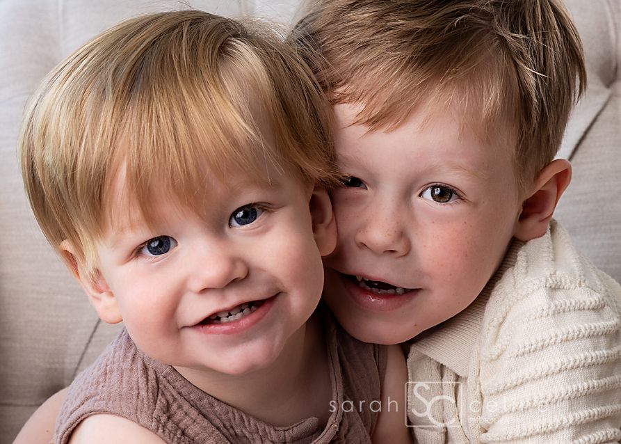 Two young children with blonde hair smile together while sitting on a beige couch.