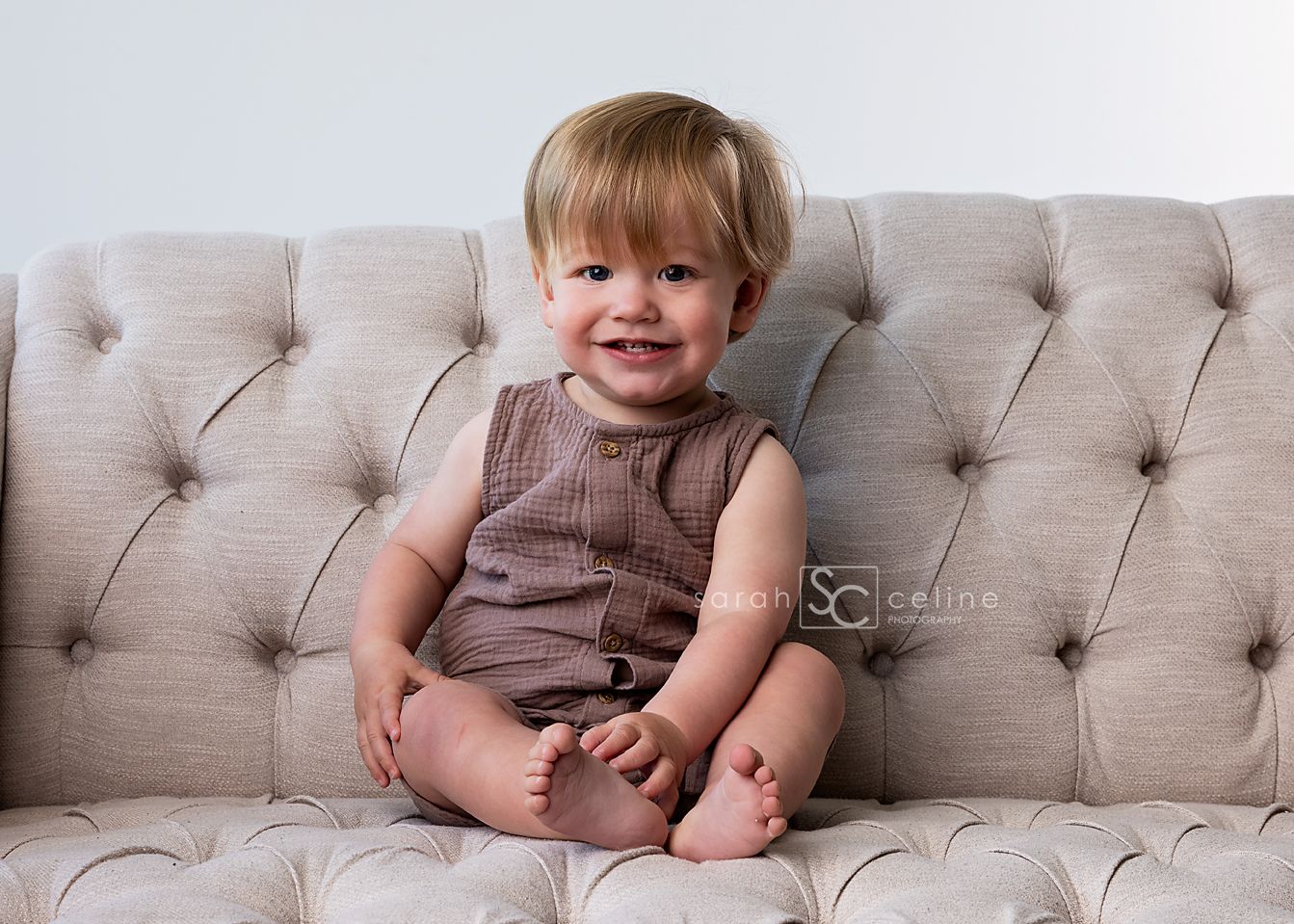 Toddler in brown romper sitting on tufted gray couch with happy expression.