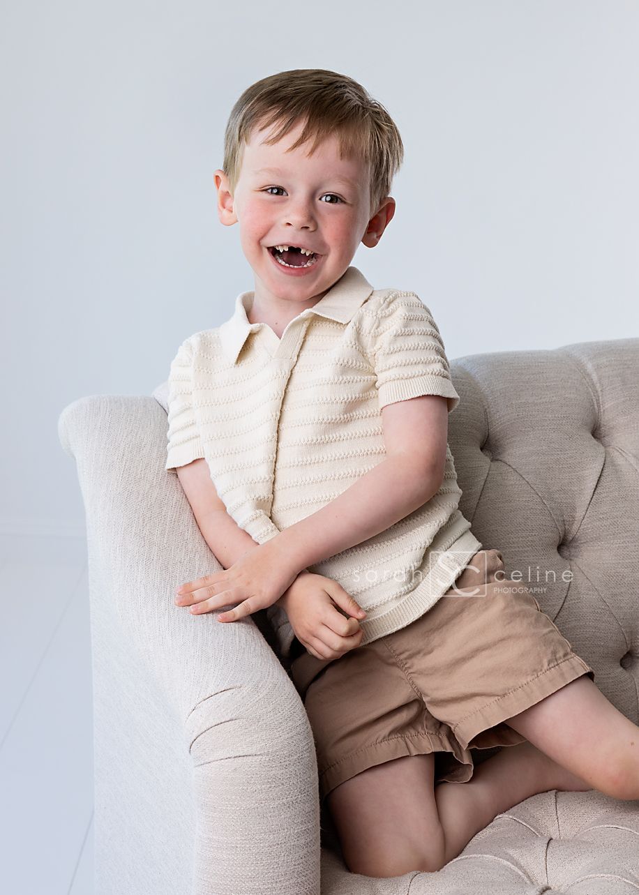 A young child wearing a cream-colored shirt and tan shorts sits casually on a light gray upholstered chair.