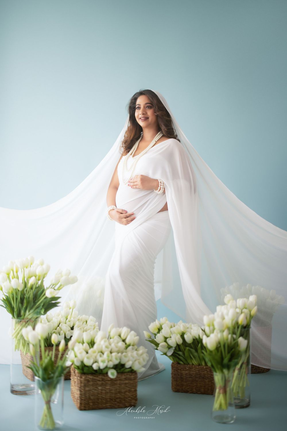 An ethereal maternity photo with flowing white fabric and white tulips arranged in baskets and vases against a blue backdrop.