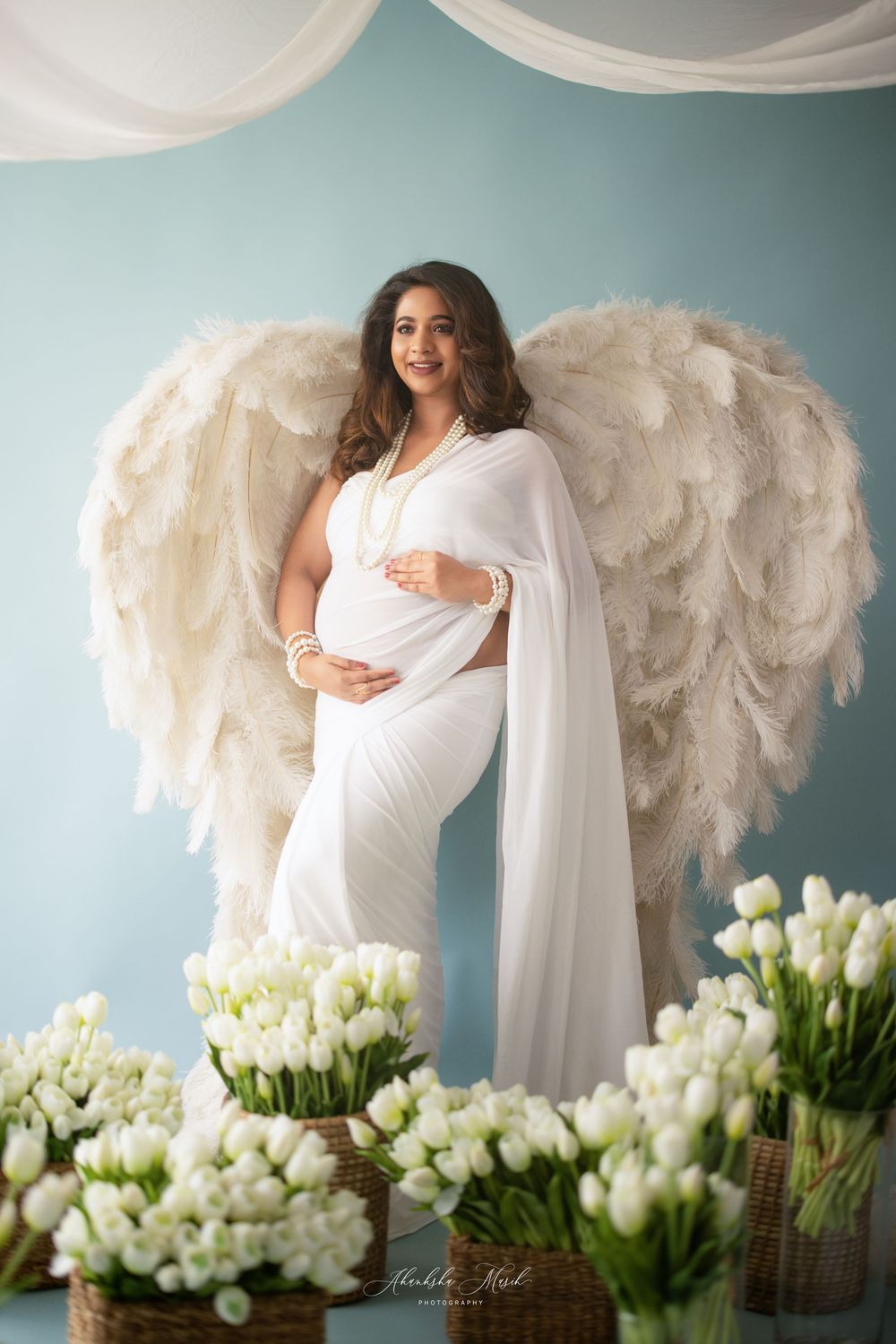 Ethereal maternity photo with large white feather wings against turquoise wall surrounded by white tulip flowers.
