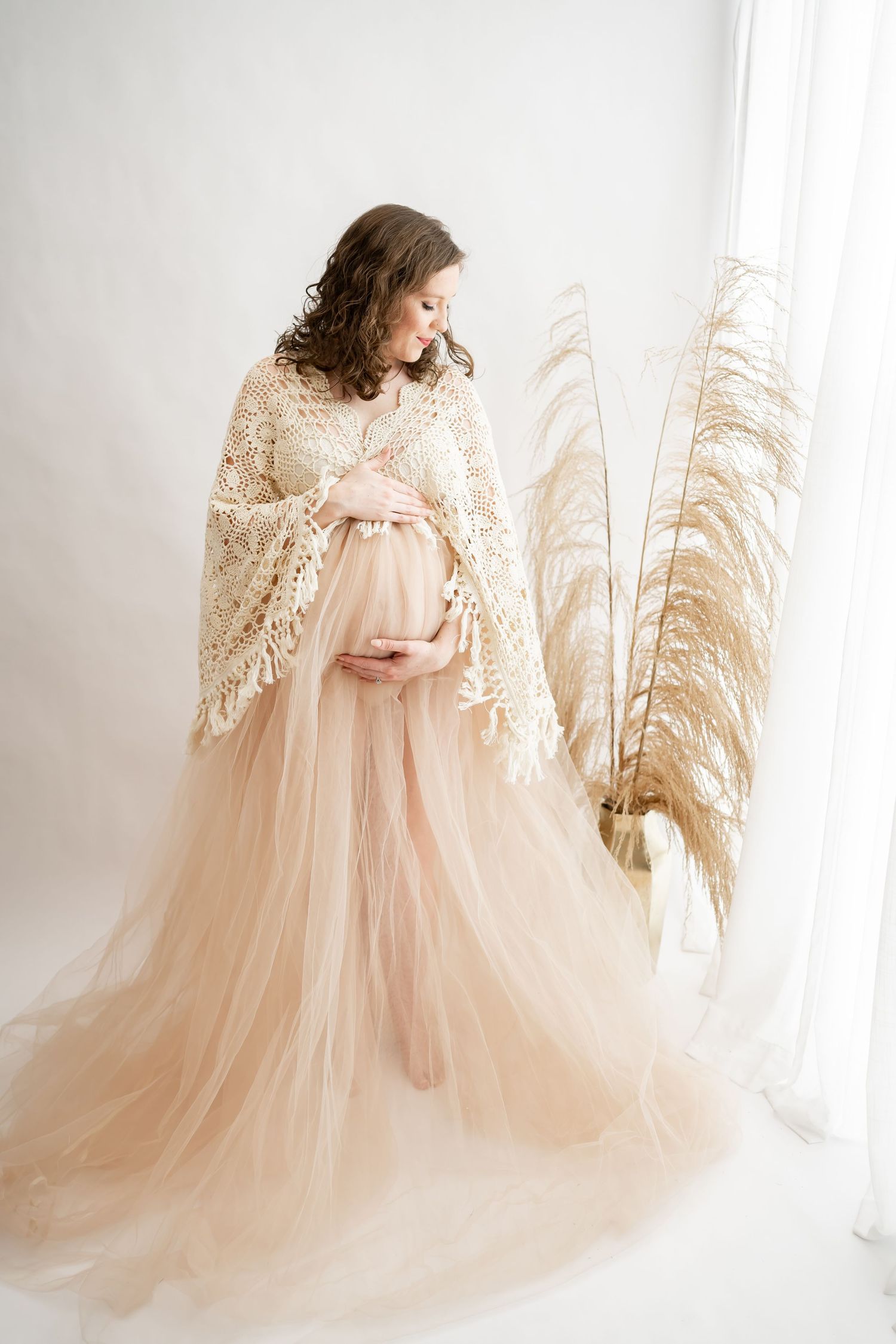 Ethereal maternity photo featuring a flowing blush tulle gown and delicate lace shawl beside pampas grass in natural light.