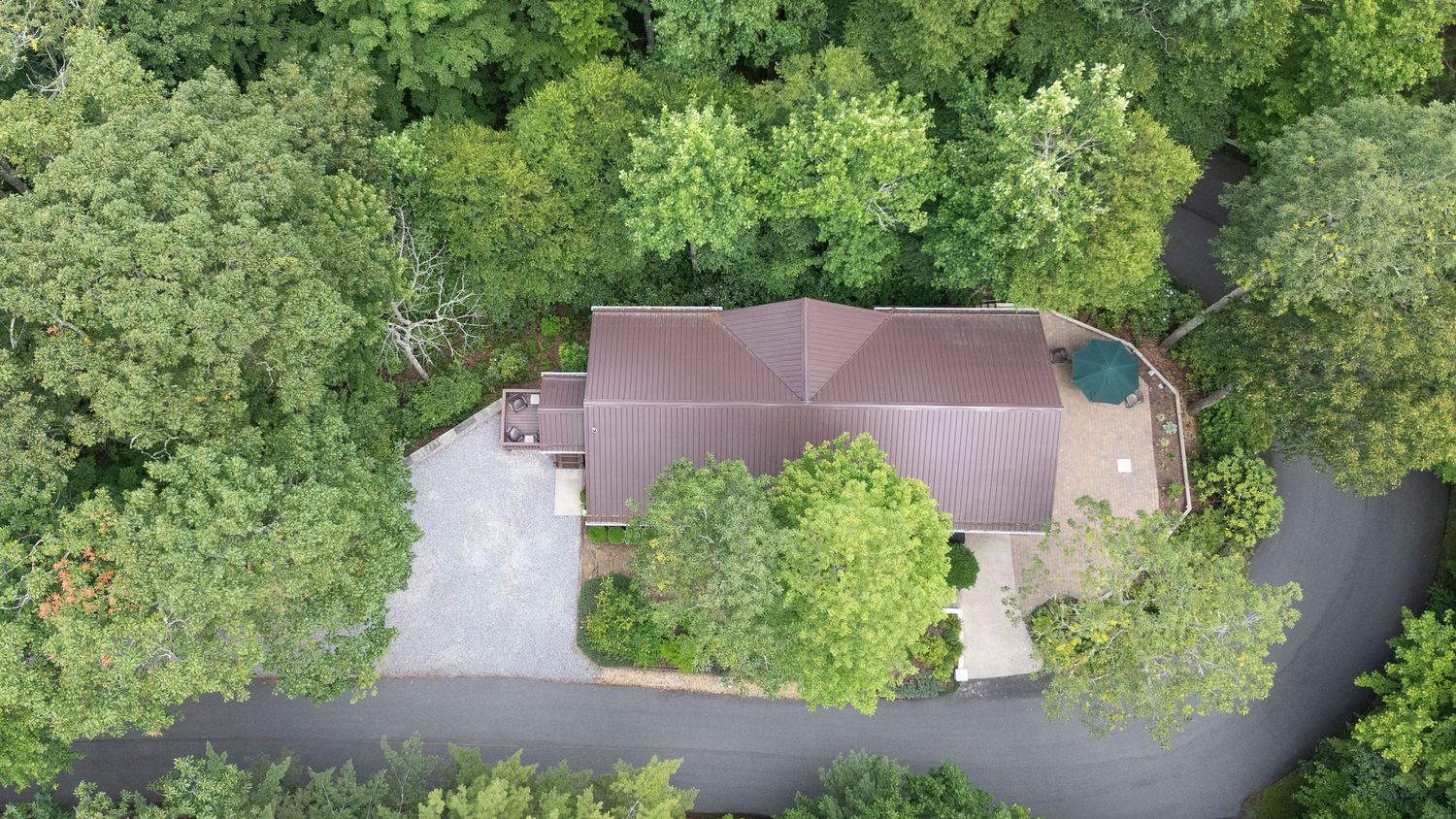 Aerial drone view of a brown house with brown roof surrounded by lush green trees and curved driveway.