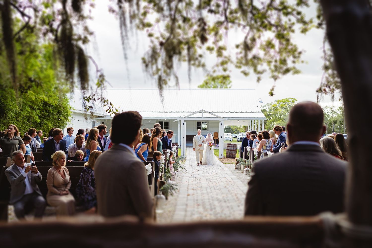 Outdoor wedding ceremony under Spanish moss with white tent and gathered guests on brick pathway.