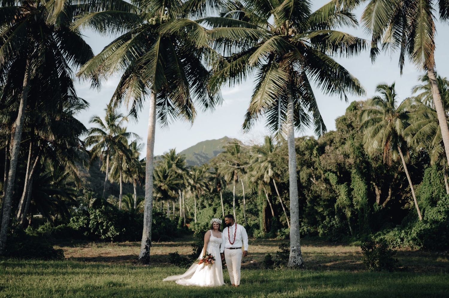 Wedding party group walks on a tropical beach with palm trees wearing flowered leis and beachwear in Hawaii.