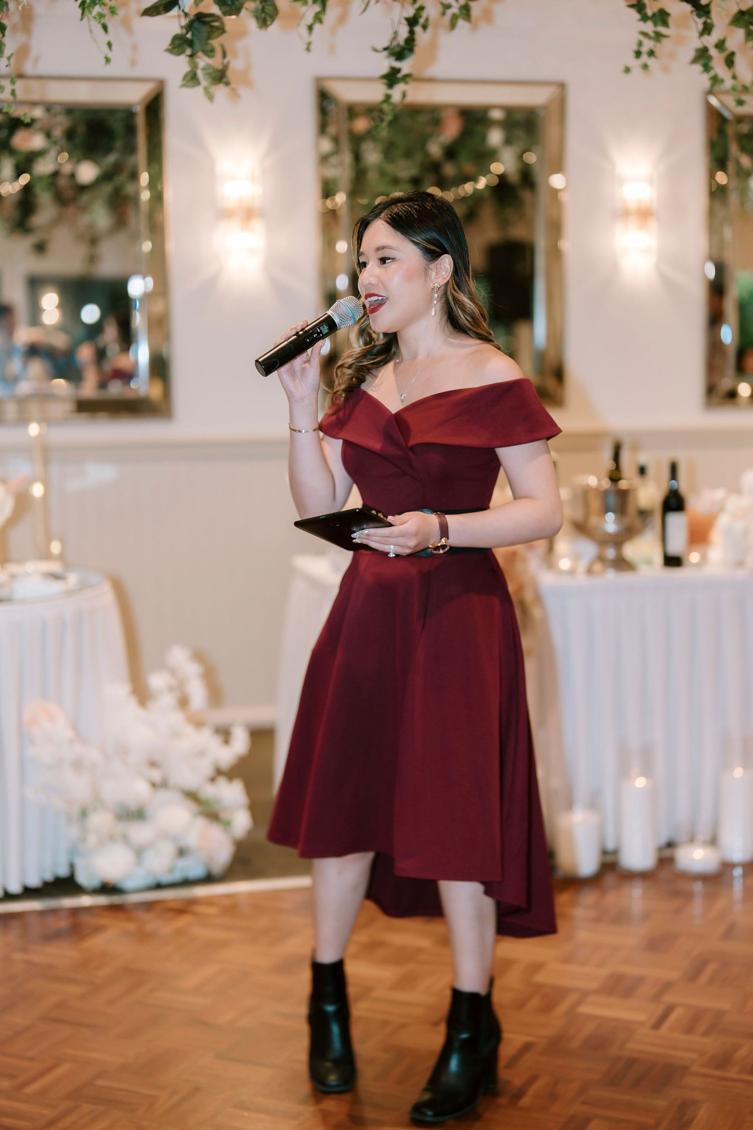 A performer in a burgundy off-shoulder dress singing with a microphone at an elegant event with candlelit tables.