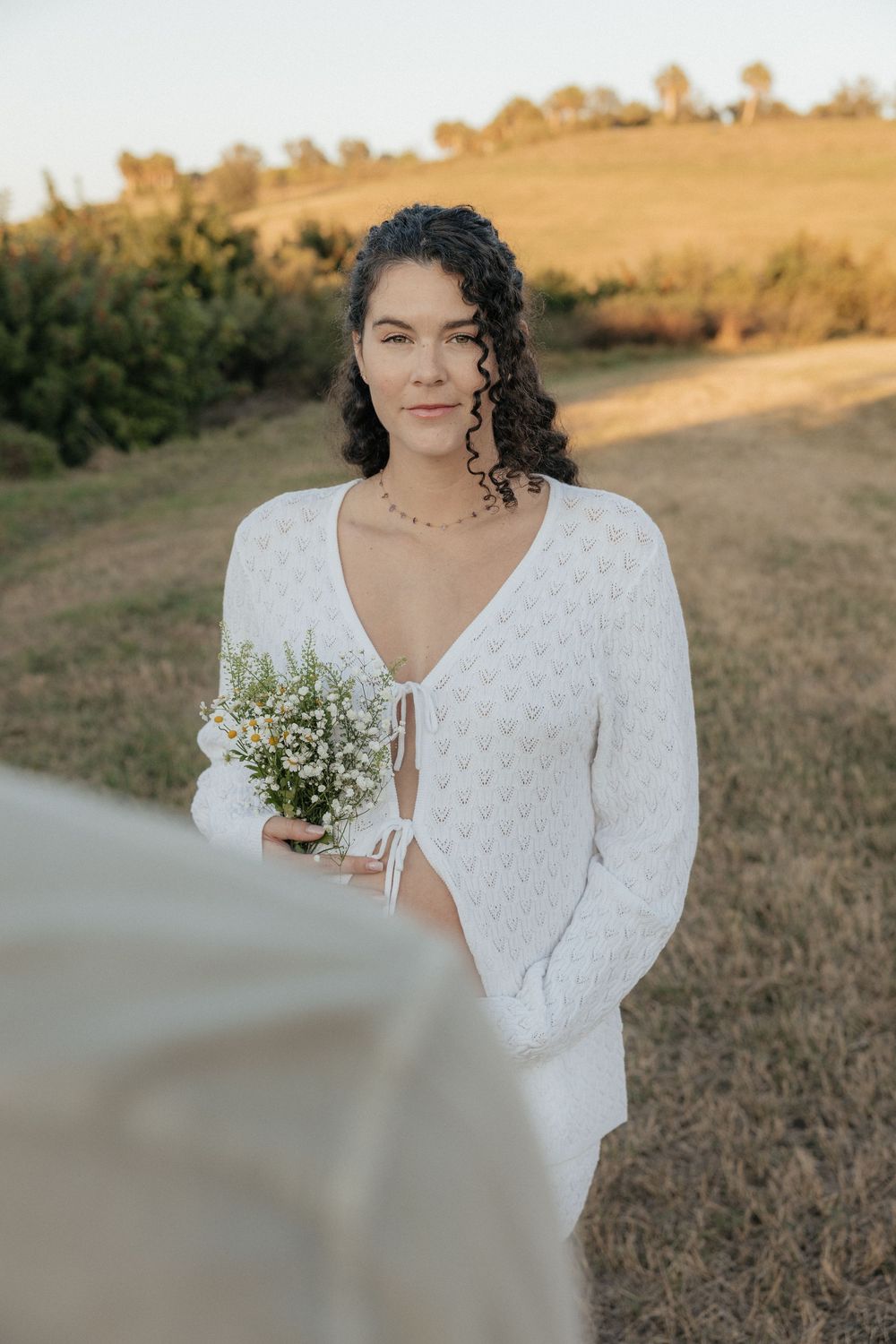 A person in a white lace dress holds delicate white baby's breath flowers in a golden outdoor setting at sunset.