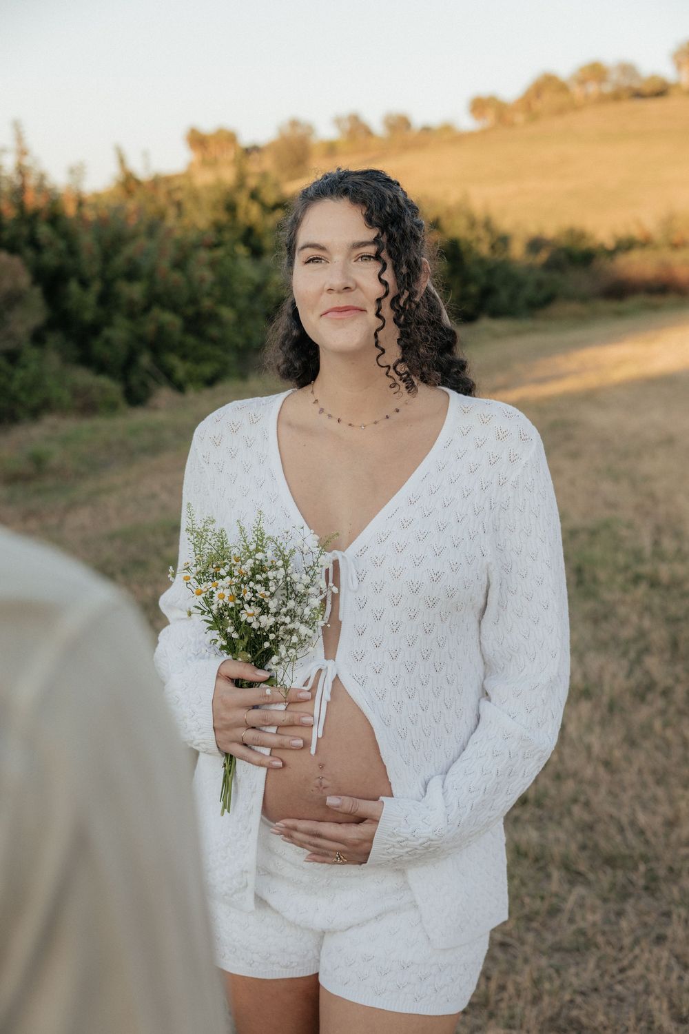 A person in a white lace romper holds a delicate bouquet of wildflowers in a sunlit outdoor setting.