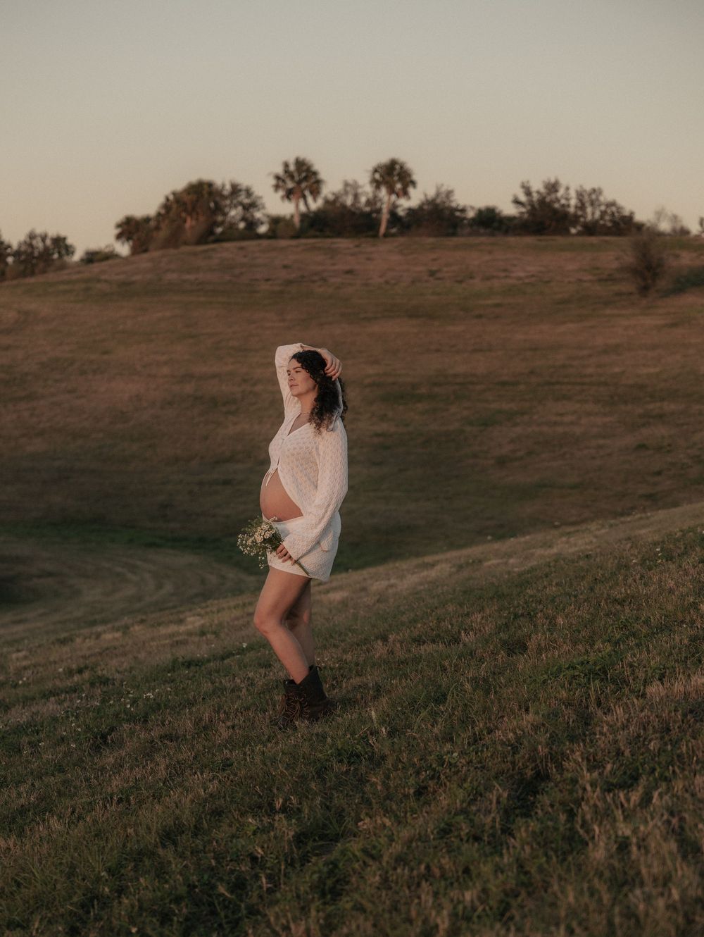 A dreamy sunset maternity photo shows a figure in white holding flowers on a hilltop.