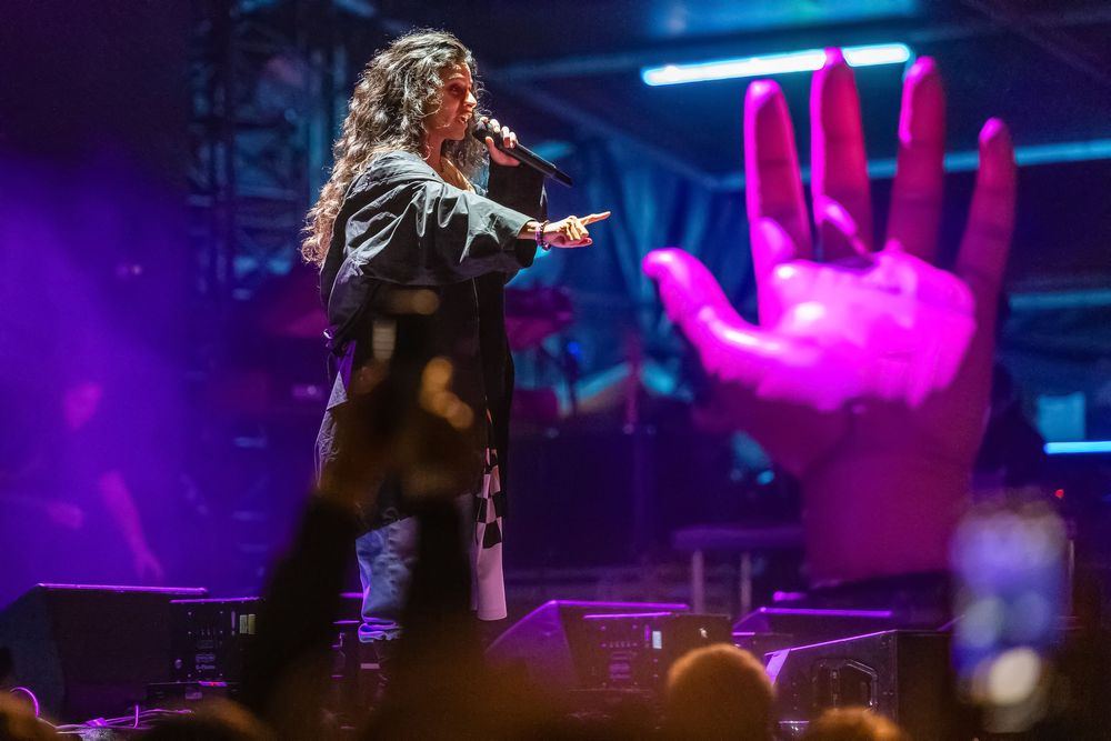 A performer on stage singing into a microphone with dramatic purple lighting and a large illuminated pink hand in the background.