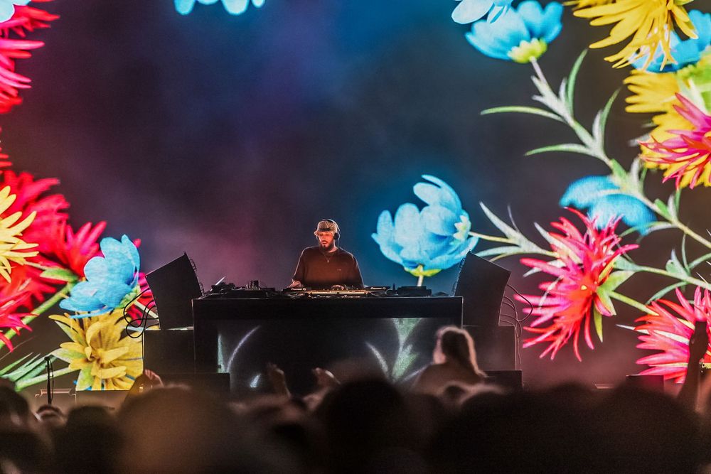 DJ performs on stage with vibrant floral visual projections and neon lighting effects during live concert performance.