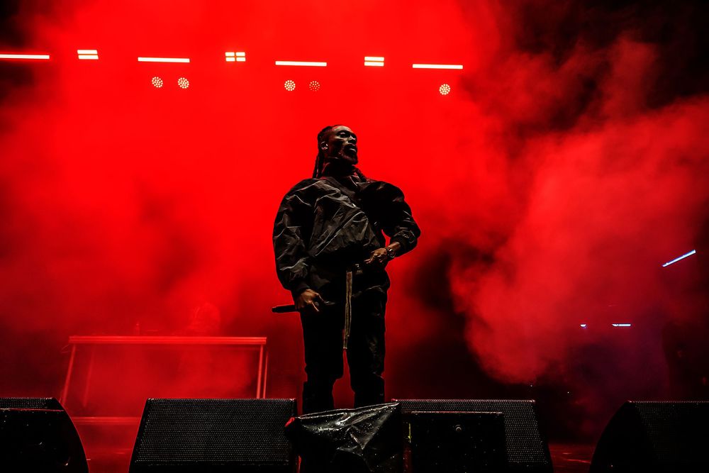 Silhouetted performer stands on stage surrounded by dramatic red smoke effects and lighting during concert performance.