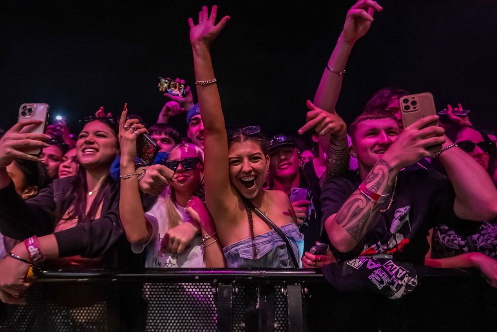 Concert crowd illuminated in purple lighting waves hands in the air while holding up phones to record the performance.