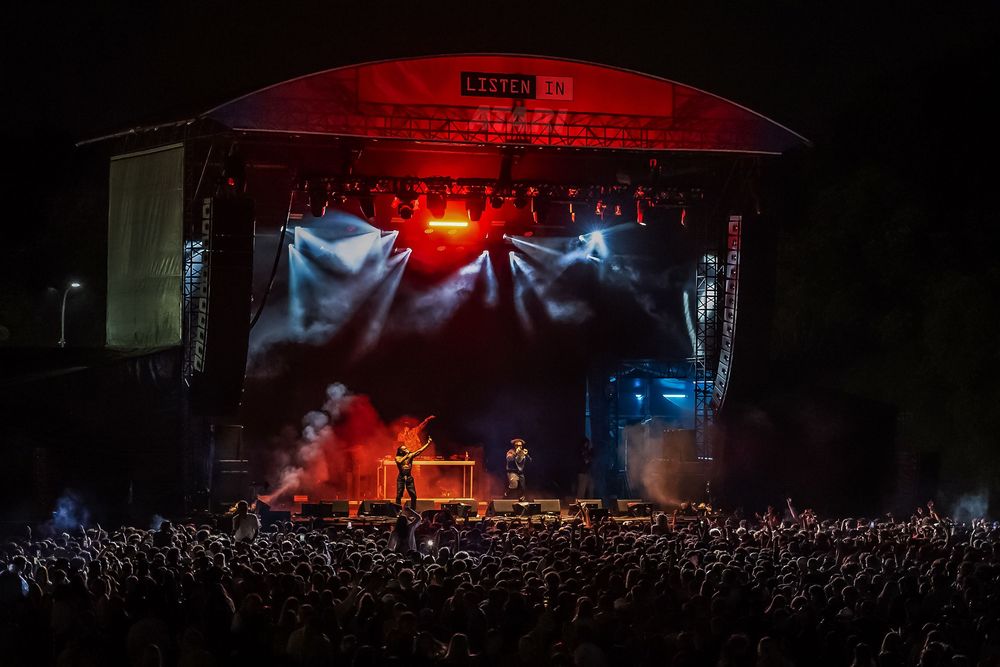 Large outdoor concert crowd at night with dramatic red lighting and smoke effects illuminating the stage and audience.