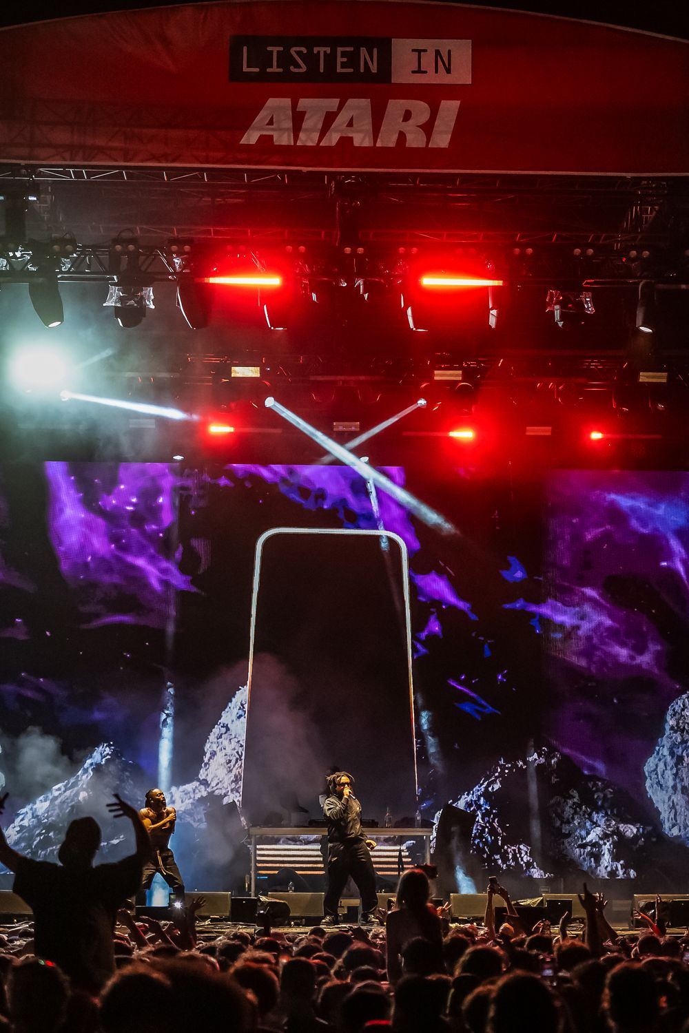 Concert stage with vibrant red and purple lighting effects showing 'LISTEN IN ATARI' signage above performers and crowd.