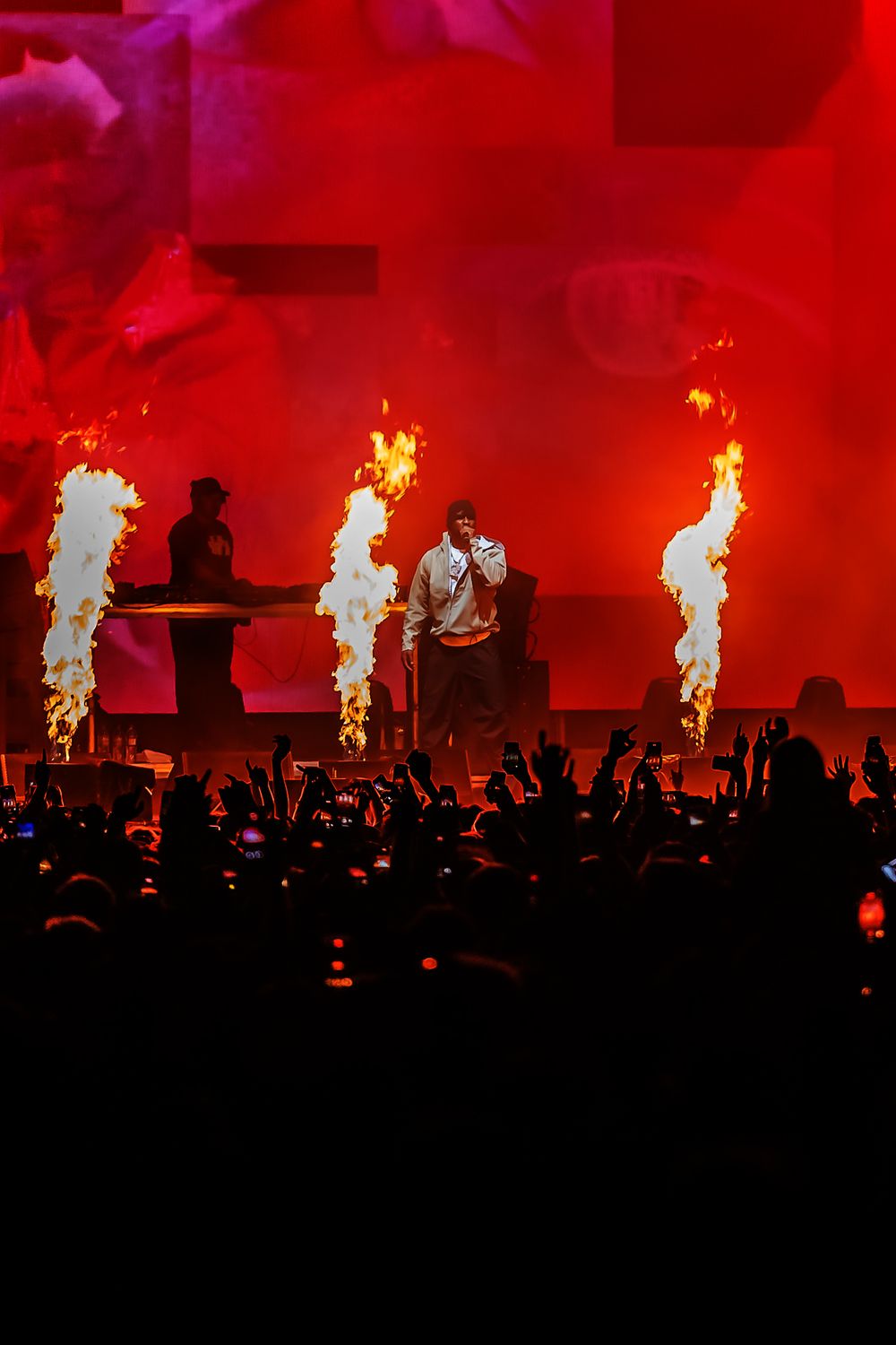 Concert stage illuminated in bright red with dramatic pyrotechnic flame effects shooting upward against a dark silhouetted crowd.