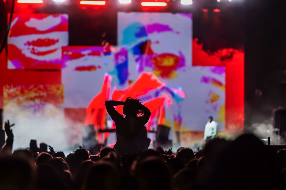 Concert crowd silhouettes against vibrant red stage lighting and video screens at a high-energy music performance.