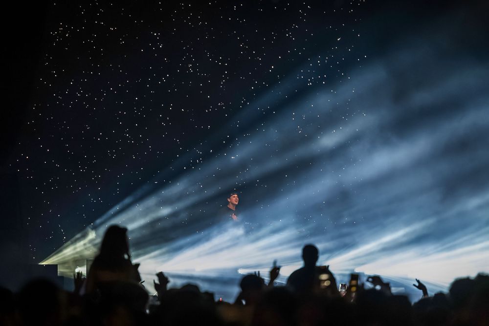 Concert crowd silhouettes against a dramatic night sky with bright spotlights creating striking beams through clouds.