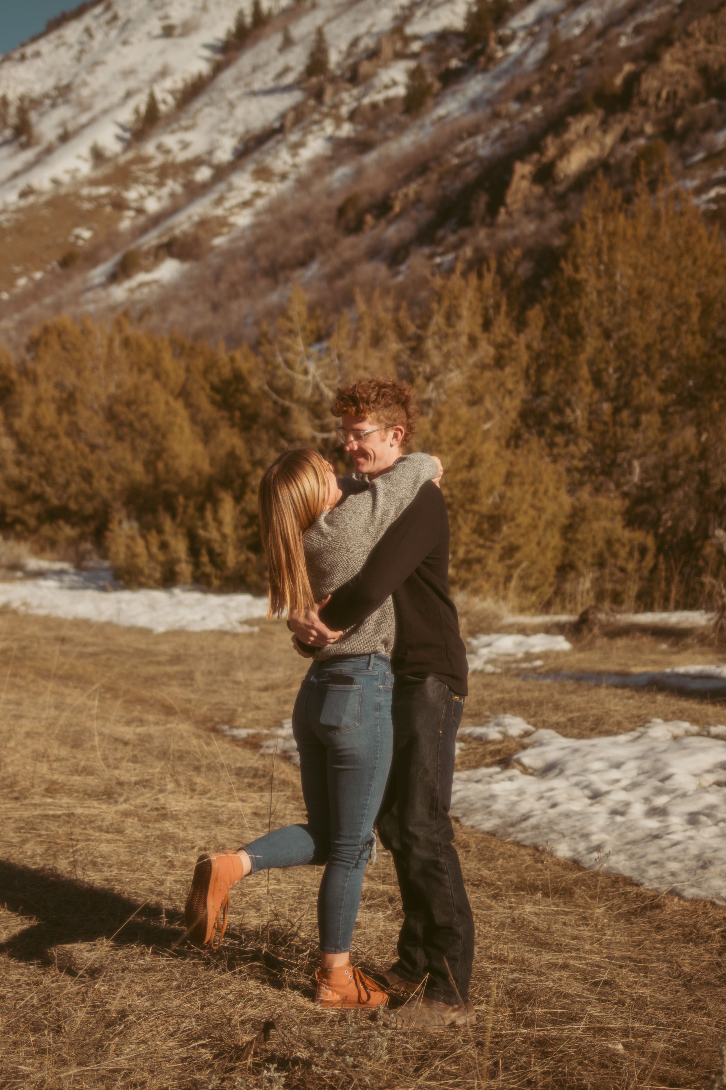 A couple embraces against snowy mountain backdrop during golden hour on a winter hillside.