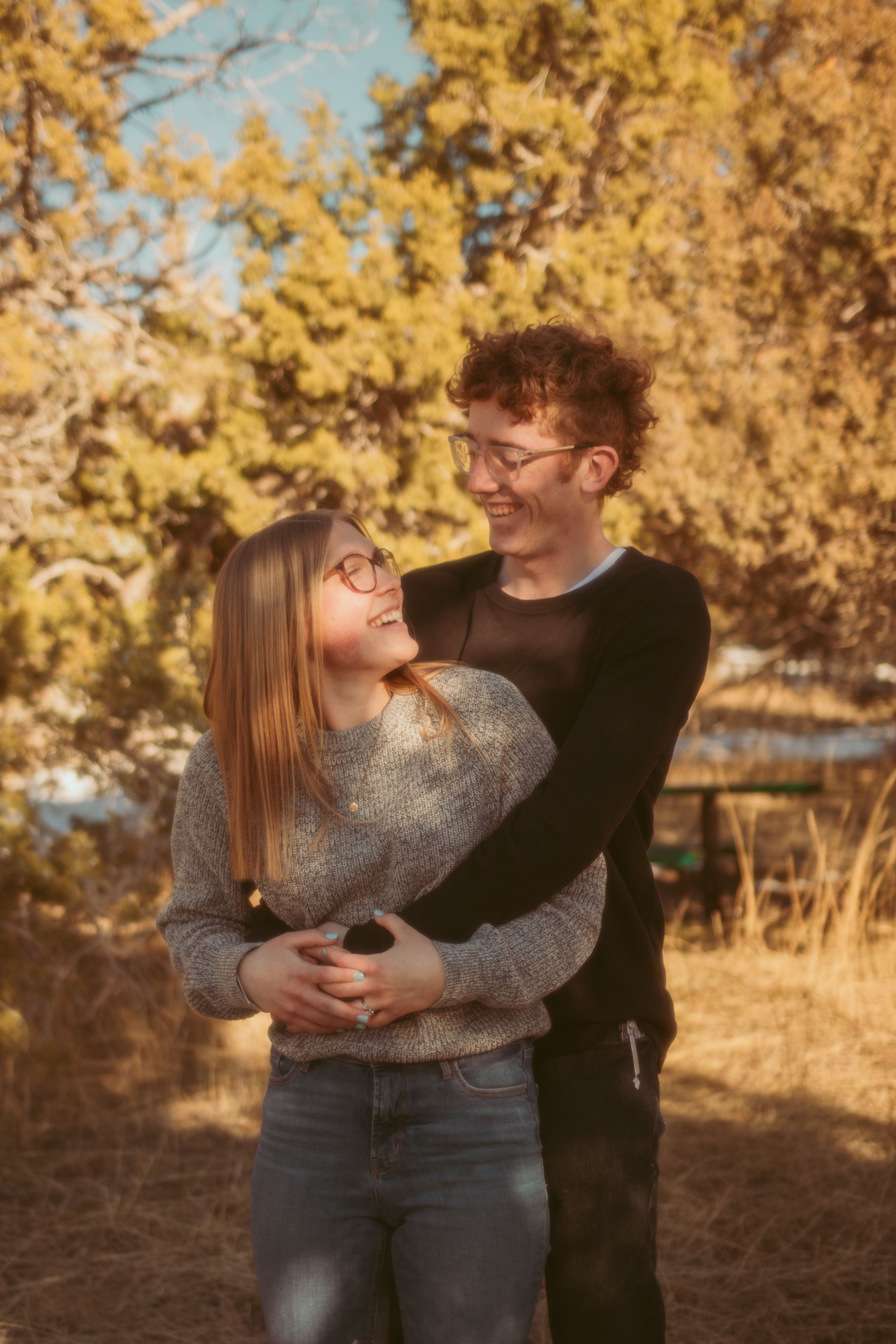 A couple wearing glasses embrace and laugh together in an autumn setting with golden foliage in the background.