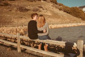 Couple shares tender moment while sitting on wooden fence overlooking scenic mountain landscape at sunset.