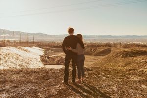 Two figures stand together watching sunset over desert landscape with mountains in background at golden hour.