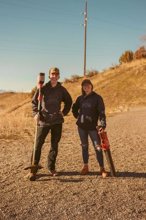 Two hikers stand on a dirt trail against a sunny hillside with power lines and golden grass in the background.