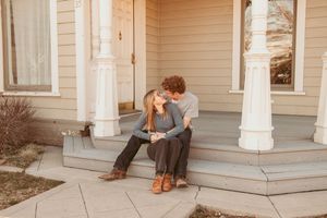 A couple sits together on the front steps of a light-colored house with white columns on the front porch.