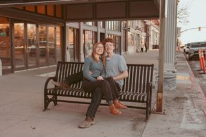 Two people sitting close together on a wooden bench outside a storefront at dusk.