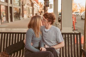 Two people share an intimate moment sitting together on a wooden bench along a city street on an autumn day.
