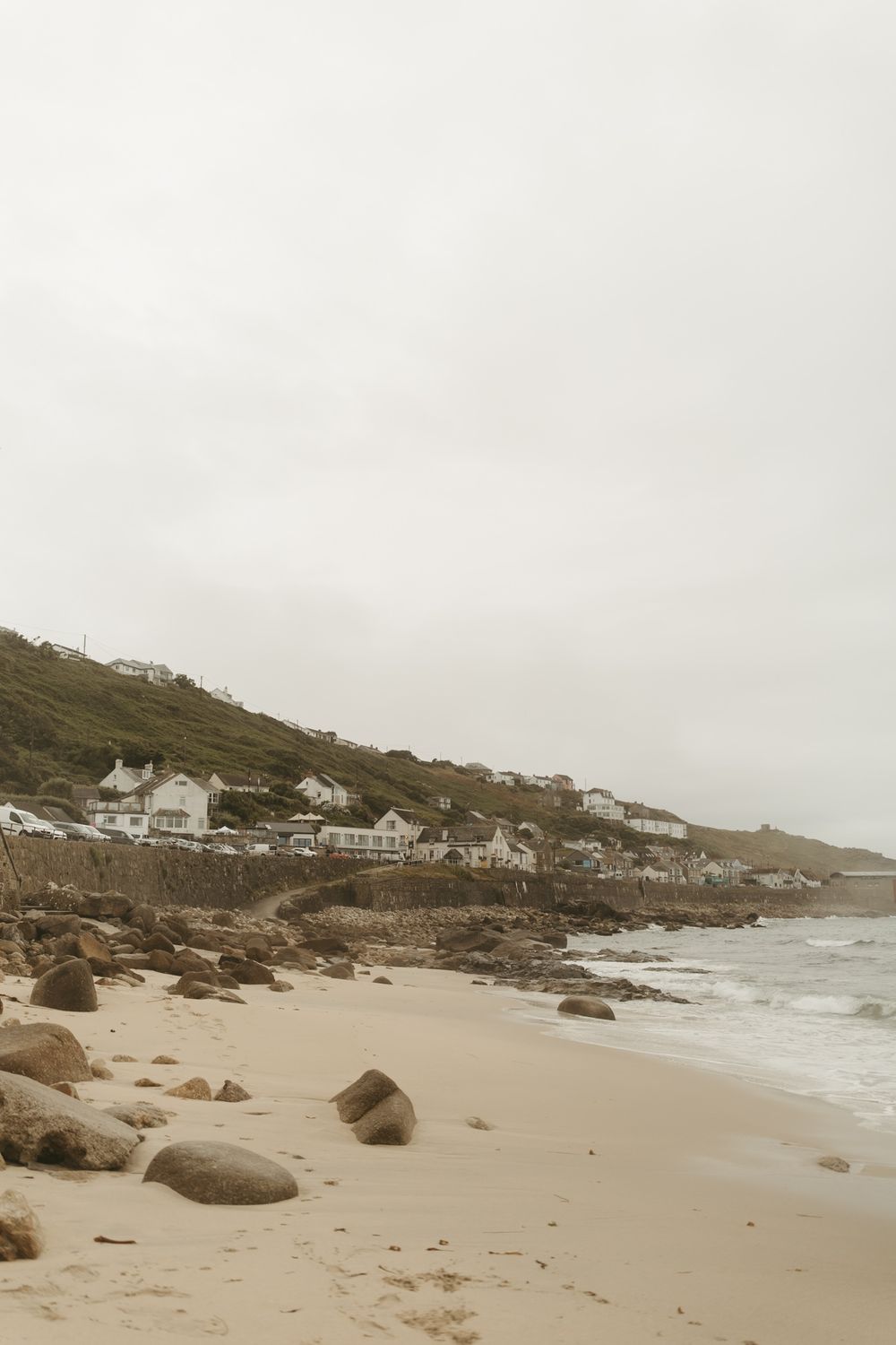 Coastal beach houses perched on rocky hillside overlooking sandy shore with scattered boulders on cloudy day.