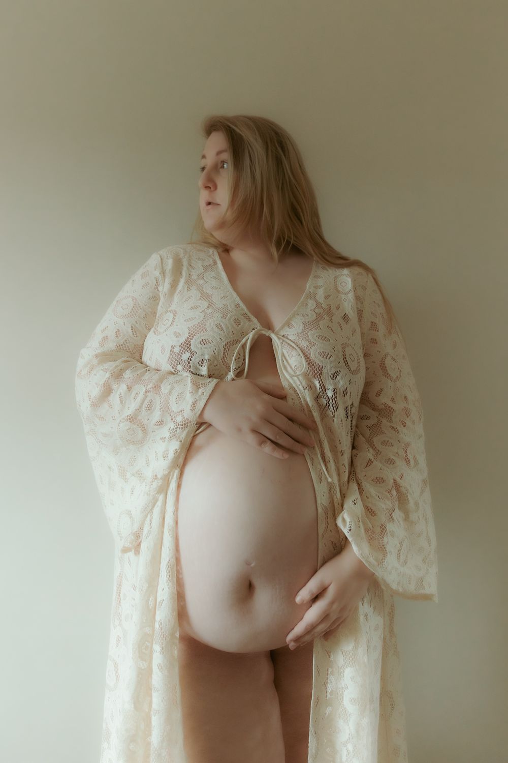 A maternity photo of an expectant mother wearing a cream lace robe against a light wall.