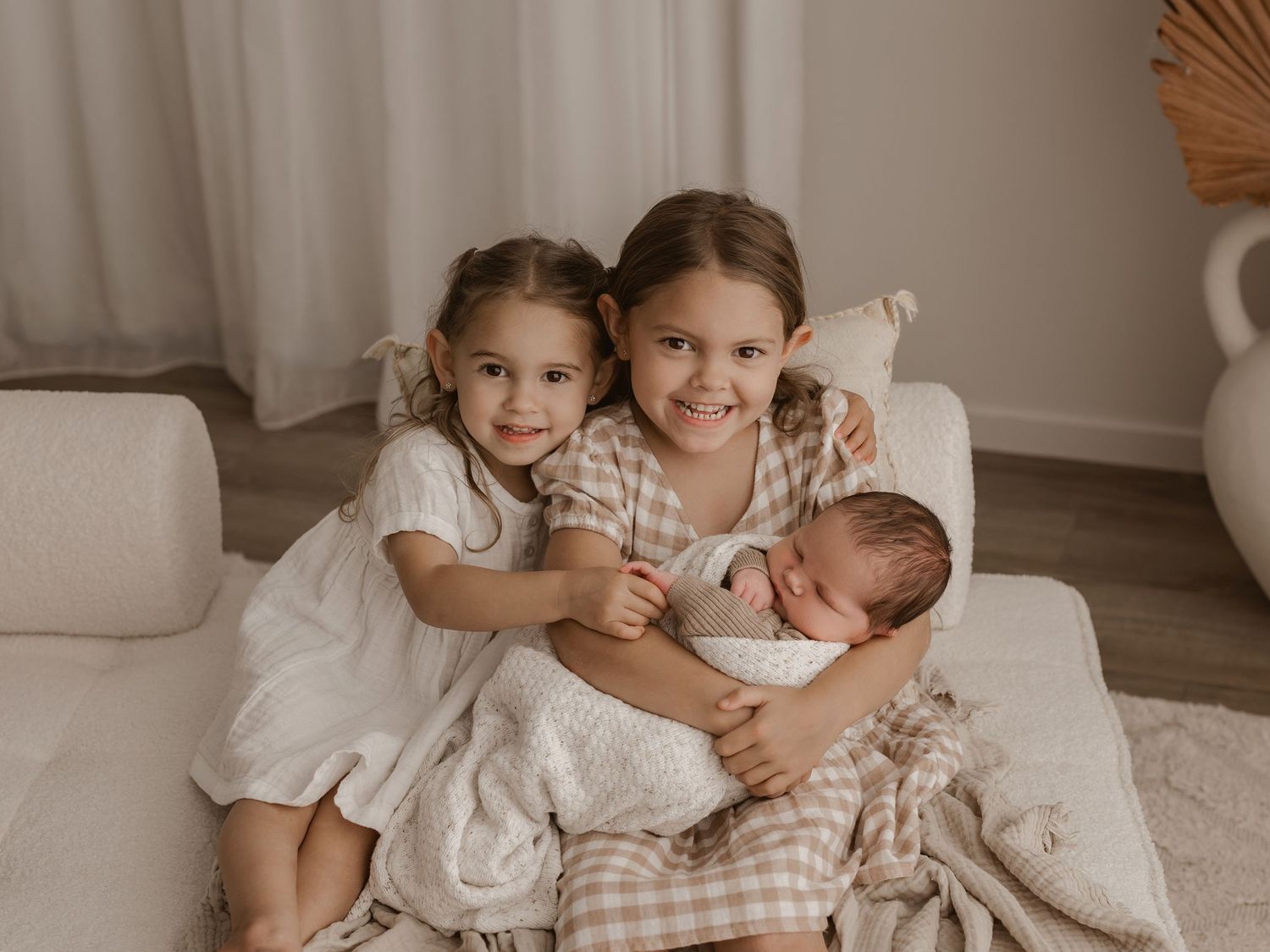 Three children in soft white and beige outfits sit close together on a cozy beige couch beside curtained windows. Photographed in a studio l