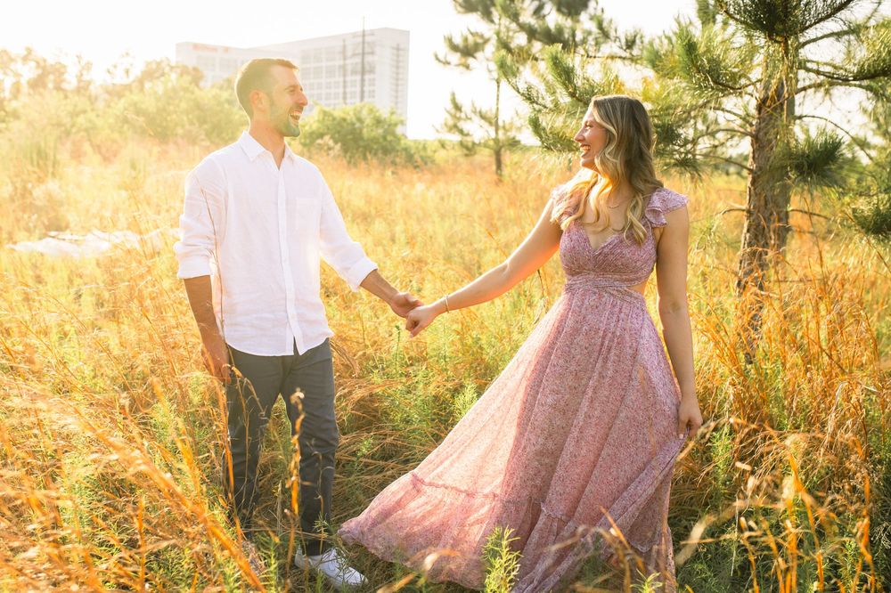 Couple shares romantic moments in golden sunlit meadow wearing pink dress and white shirt during sunset photoshoot.