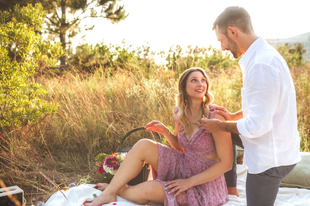 Two people sitting together in golden sunlight on grassy coastal cliff at sunset sharing intimate moment.